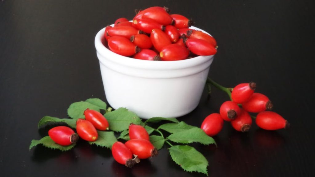 Vibrant red rose hips in a ceramic bowl atop a dark wooden table, emphasizing freshness and natural produce.